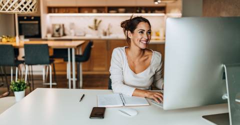 Een vrolijke vrouw die thuis aan het werk is achter de computer met op de achtergrond de keuken en eettafel